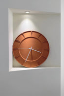 A modern round wall clock with a matte copper-orange finish and white hands, mounted on a white wall in a recessed niche with a spotlight above, Obroi