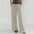 A person wearing loose, wide-leg striped pants with a vertical stripe pattern in beige and gray, paired with a dark brown top and black sandals, shown from the waist down against a plain light background, Obroi