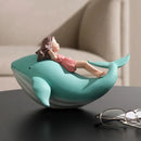 A decorative whale-shaped object in mint green and white, with a small reclining girl figurine inside, placed on a dark tabletop in a softly lit indoor setting. A pair of eyeglasses is partially visible in the foreground, Obroi