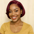 A smiling woman with a short, glossy, deep burgundy bob wig, wearing a mustard yellow top and a delicate gold necklace, set against a plain beige background, Obroi