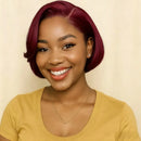 A smiling woman with a short, glossy, deep burgundy bob wig, wearing a mustard yellow top and a delicate gold necklace, set against a plain beige background, Obroi