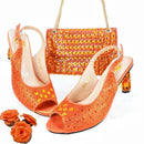A matching women's accessory set featuring a pair of orange rhinestone-embellished slingback peep-toe heels and a matching orange rhinestone-embellished handbag with gold chain straps, displayed on a white background with decorative orange roses in the corner, Obroi