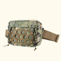 A tactical waist bag with a green and brown camouflage pattern, featuring a tan honeycomb-patterned front panel and an adjustable tan webbing strap, shown against a solid cream background, Obroi