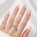 A close-up of a hand with long, glossy almond-shaped nails decorated with pink and white floral designs, wearing a silver twisted ring on a soft white background, Obroi