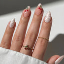 Hand with almond-shaped nails painted in white and pink, featuring red cherry designs, wearing two rings on a light background, Obroi