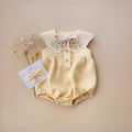 Yellow baby romper with floral collar and matching bow on a beige background, Obroi