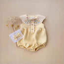 Yellow baby romper with floral collar and matching bow on a beige background, Obroi