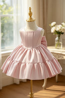 A light pink girls' party dress displayed on a cream dress form with a gold finial, set in a softly lit room with sheer curtains, a window, and a table with white flowers in the background, Obroi