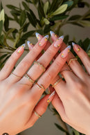 Close-up of hands with decorative nail art and gold rings against a blurred green plant background, Obroi