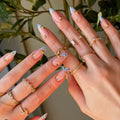 Close-up of hands with decorative nail art and gold rings against a blurred green plant background, Obroi