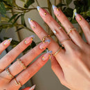 Close-up of hands with decorative nail art and gold rings against a blurred green plant background, Obroi