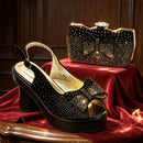 A black peep-toe platform sandal with a rhinestone-embellished bow and heel, displayed on a red velvet surface next to a matching rhinestone clutch purse, Obroi