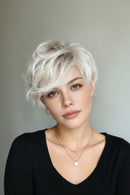 A woman with a short, textured platinum blonde wig, wearing a black top and delicate gold necklaces, set against a plain gray background, Obroi