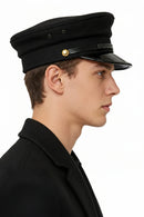 A man in profile wearing a black military-style peaked cap with a glossy black visor and a small gold button, against a plain white background, Obroi