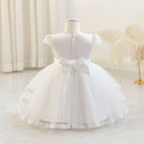 White toddler girl's party dress displayed on a dress form mannequin, featuring a tiered tulle skirt, lace trim, and a large bow at the back waist, Obroi