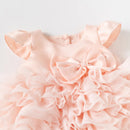 A close-up of a light pink girls' dress with ruffled fabric, flutter sleeves, and a large bow at the neckline, shown against a plain light background, Obroi