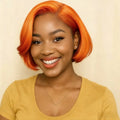 A smiling woman with a bright orange short bob wig, wearing a mustard yellow top and a delicate gold necklace, set against a plain beige background, Obroi