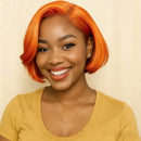 A smiling woman with a bright orange short bob wig, wearing a mustard yellow top and a delicate gold necklace, set against a plain beige background, Obroi