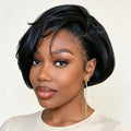 A woman with a short, styled black wig or hairpiece shown from the shoulders up against a plain light beige background. The hair is glossy and neatly cut with soft side-swept bangs, and she is wearing a light beige top and a dangling gold earring, Obroi
