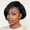 A woman with a short, styled black wig or hairpiece shown from the shoulders up against a plain light beige background. The hair is glossy and neatly cut with soft side-swept bangs, and she is wearing a light beige top and a dangling gold earring, Obroi