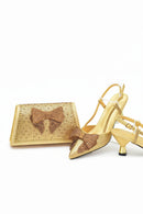 A gold metallic shoe and matching clutch set, both covered in rhinestones and featuring a large bow detail on the front, displayed on a clean white background, Obroi
