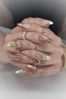 A close-up of a hand showcasing a detailed artificial nail set with long almond-shaped nails. The nails feature a glossy nude-pink base with white tips, gold metallic leaf designs, and small pearl accents, Obroi