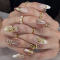 A close-up of a hand showcasing a detailed artificial nail set with long almond-shaped nails. The nails feature a glossy nude-pink base with white tips, gold metallic leaf designs, and small pearl accents, Obroi