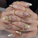 A close-up of a hand showcasing a detailed artificial nail set with long almond-shaped nails. The nails feature a glossy nude-pink base with white tips, gold metallic leaf designs, and small pearl accents, Obroi