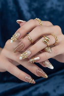 A close-up of a hand showcasing a detailed artificial nail set with long almond-shaped nails. The nails feature a glossy nude-pink base with white tips, gold metallic leaf designs, and small pearl accents, Obroi