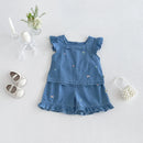 A baby girl's two-piece denim outfit in medium blue, featuring a ruffle-sleeve top and matching ruffled shorts with small embroidered butterfly details. The outfit is styled in a flat-lay arrangement on a light gray background, accompanied by baby shoes, a small pearl headband, and a glass vase with flowers, Obroi