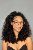 A three-panel collage showing a woman modeling a voluminous, curly black wig. The left panel shows a front-facing view of the wig with large brown-framed glasses, and the right panels show side and back views of the wig against a neutral gray background, Obroi