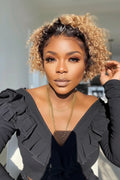 A close-up portrait of a woman with short, curly hair in a warm blonde and dark brown blend, wearing a black ruffled top and gold necklaces, Obroi