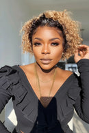 A close-up portrait of a woman with short, curly hair in a warm blonde and dark brown blend, wearing a black ruffled top and gold necklaces, Obroi