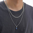 Person wearing a silver chain necklace with a blue pendant on a plain background, Obroi