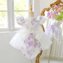 A white girls' party dress with a purple and lavender floral print, displayed on a dress form. The dress features puff sleeves and a large matching bow on the back, Obroi