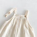 A close-up of a cream-colored crinkle gauze baby dress with shoulder straps and decorative wooden buttons, laid flat against a textured white background, Obroi