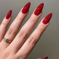 A close-up of a hand with long, almond-shaped nails painted in a sparkling deep red glitter polish, wearing a gold ring with a clear rectangular gemstone against a soft gray background, Obroi
