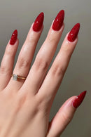 A close-up of a hand with long, almond-shaped nails painted in a sparkling deep red glitter polish, wearing a gold ring with a clear rectangular gemstone against a soft gray background, Obroi