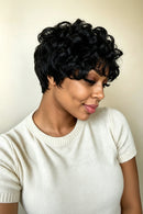 A woman wearing a short, curly black wig and a cream-colored knit top, shown in a close-up portrait against a plain beige background, Obroi