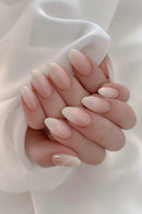 A close-up of a hand with long, almond-shaped press-on nails in a glossy nude pink color, resting against a soft white fabric background, Obroi