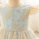 A close-up of a child's dress on a mannequin, featuring light blue lace with gold floral embroidery, ruffled cap sleeves, and a waistband decorated with a row of white faux pearls, Obroi