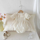 White lace baby dress on a hanger with white shoes and a vase of flowers on a white surface, Obroi