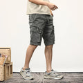 Person wearing gray cargo shorts standing on a wooden floor with a white wall background, Obroi