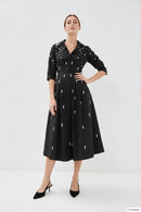 A model wears a black midi shirt dress with three-quarter sleeves, a button-up front, and pockets, decorated with small white embroidered motifs. The dress is styled with black pointed-toe heels against a plain white studio background, Obroi