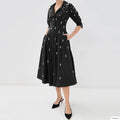 A model wears a black midi shirt dress with three-quarter sleeves, a button-up front, and pockets, decorated with small white embroidered motifs. The dress is styled with black pointed-toe heels against a plain white studio background, Obroi