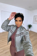 A woman wearing a sage green bomber jacket with brown trim over a mauve ribbed top, posing in a bright room with light wood floors, Obroi