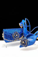 A coordinated blue sequined fashion set featuring a clutch bag, pointed shoe, and high-heeled sandal, each decorated with a large gold-tone beaded flower and chain detail, displayed against a black background, Obroi