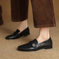Black loafers worn with brown pants on a beige surface, Obroi