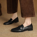 Black loafers worn with brown pants on a beige surface, Obroi