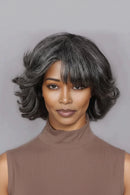 A woman wearing a short, wavy wig with dark gray and black tones, styled with a straight fringe, against a solid gray background, Obroi